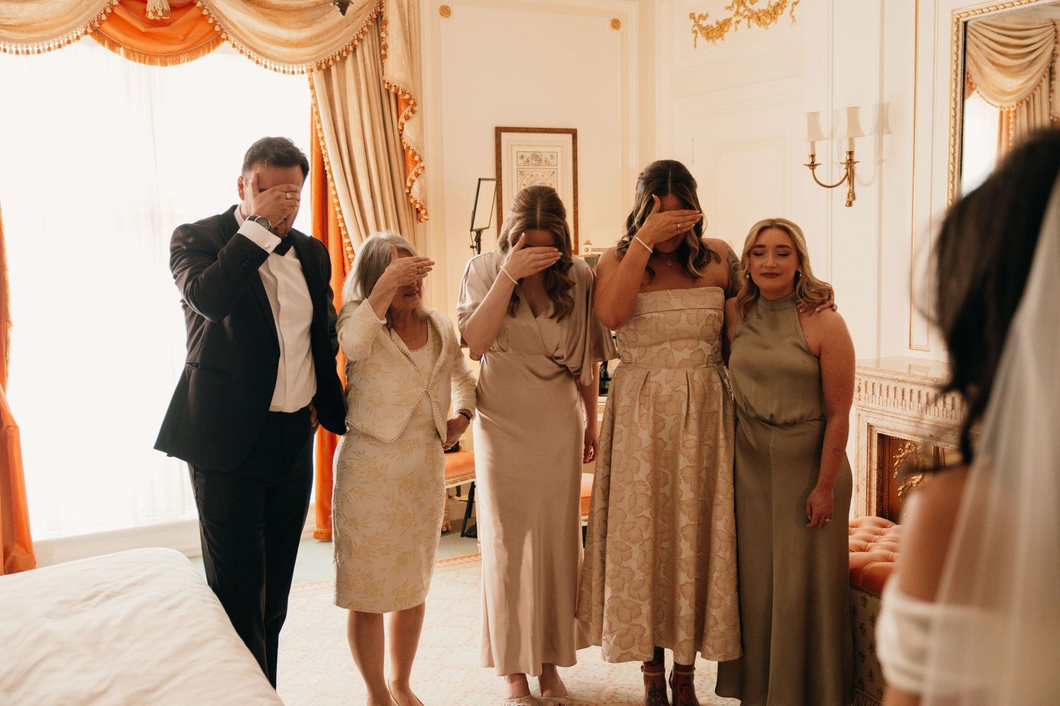 Emotional bridesmaids and family members reacting with tears as bride reveals her wedding dress in hotel room.