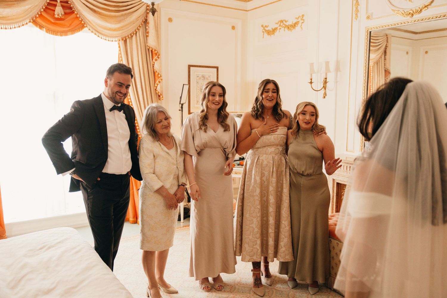 Family members react with joy as bride reveals her wedding dress in an elegant room.
