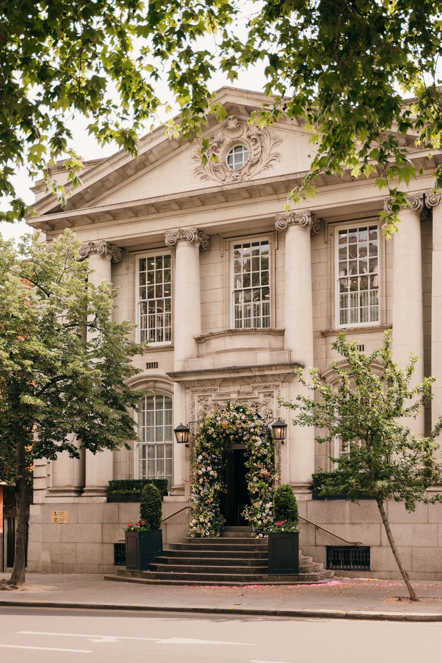 Elegant neoclassical wedding venue exterior with stone columns, floral entrance arch, and tree-lined street.