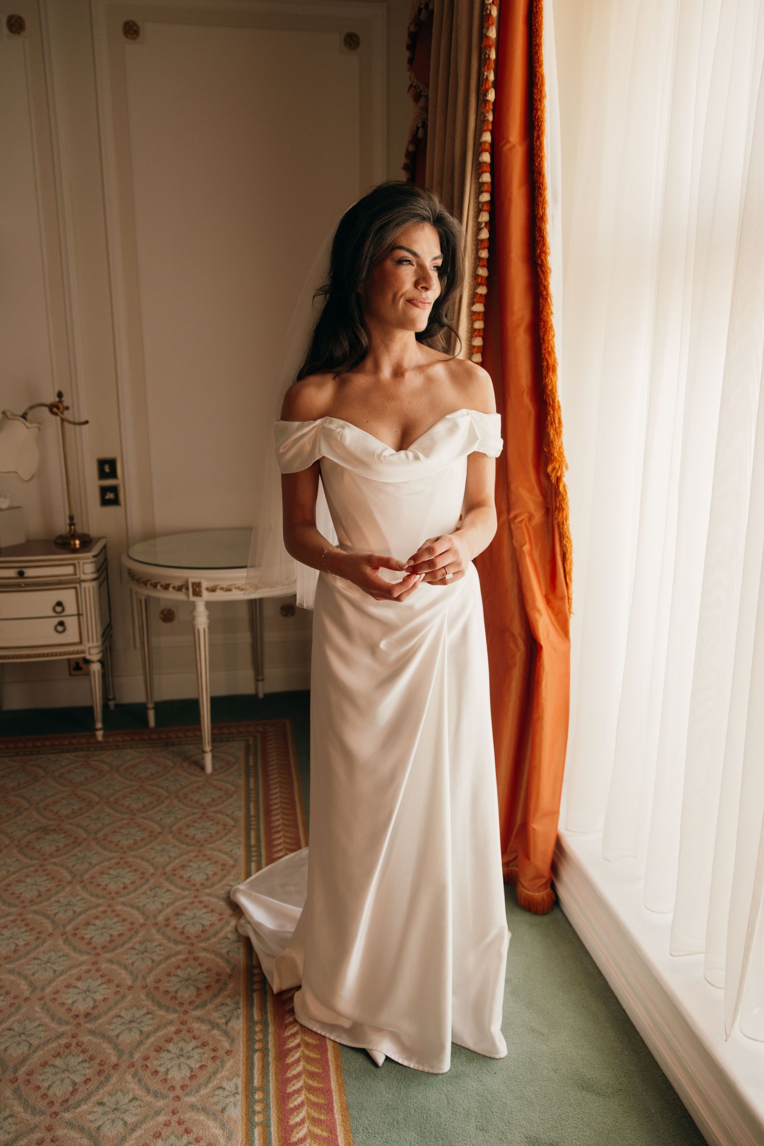 Bride in off-shoulder white satin wedding gown standing by a sunlit window in an elegant room.