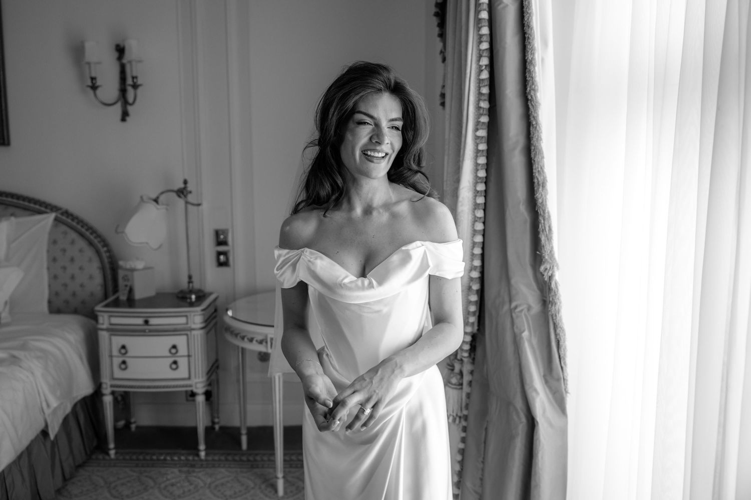 Radiant bride in off-shoulder white satin gown smiles joyfully by window in elegant hotel room.