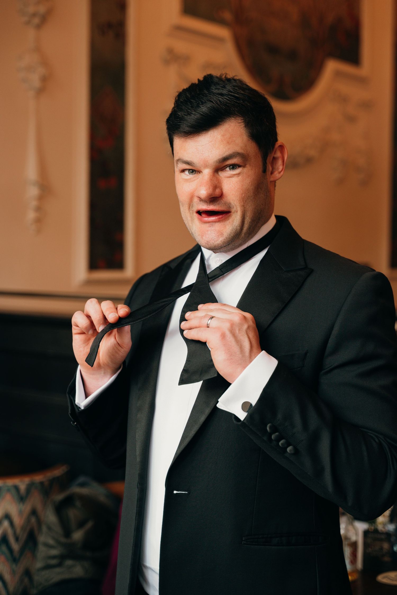 Groom in black tuxedo adjusting his bow tie in an elegant ornate room before the wedding ceremony.
