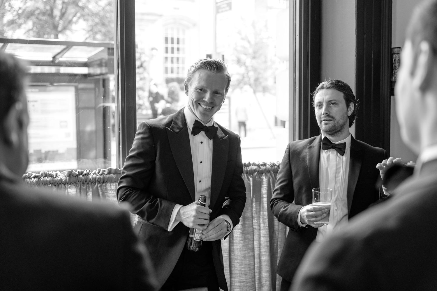 Two groomsmen in tuxedos smile and chat during a wedding reception in black and white.
