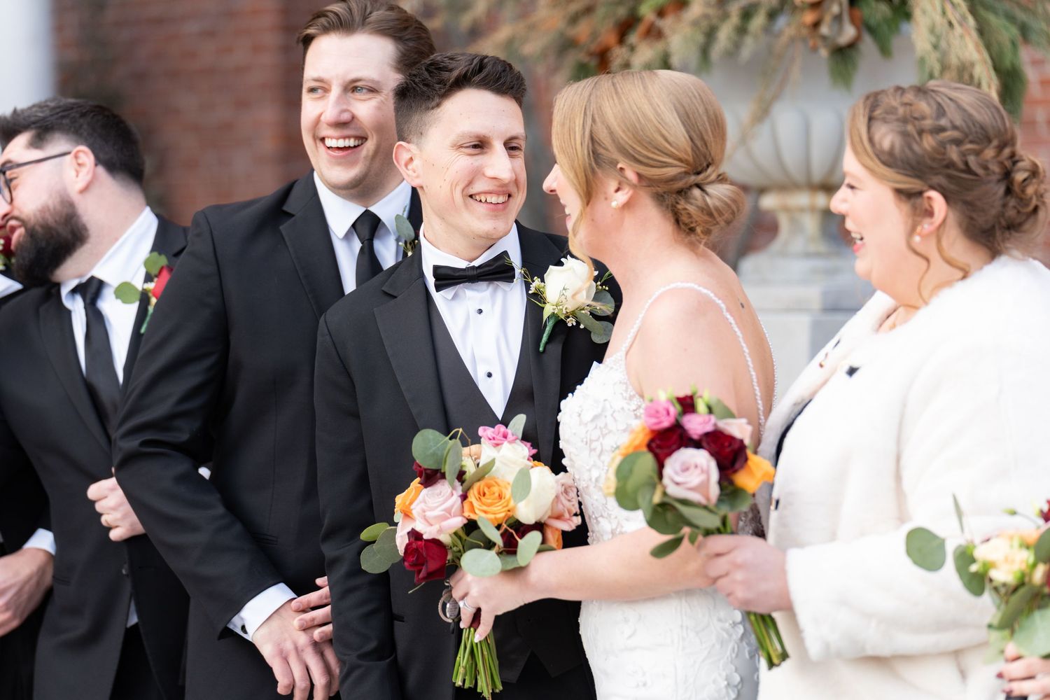 Bride and groom laughing with wedding party outdoors, bride holding colorful bouquet of roses in elegant white gown.