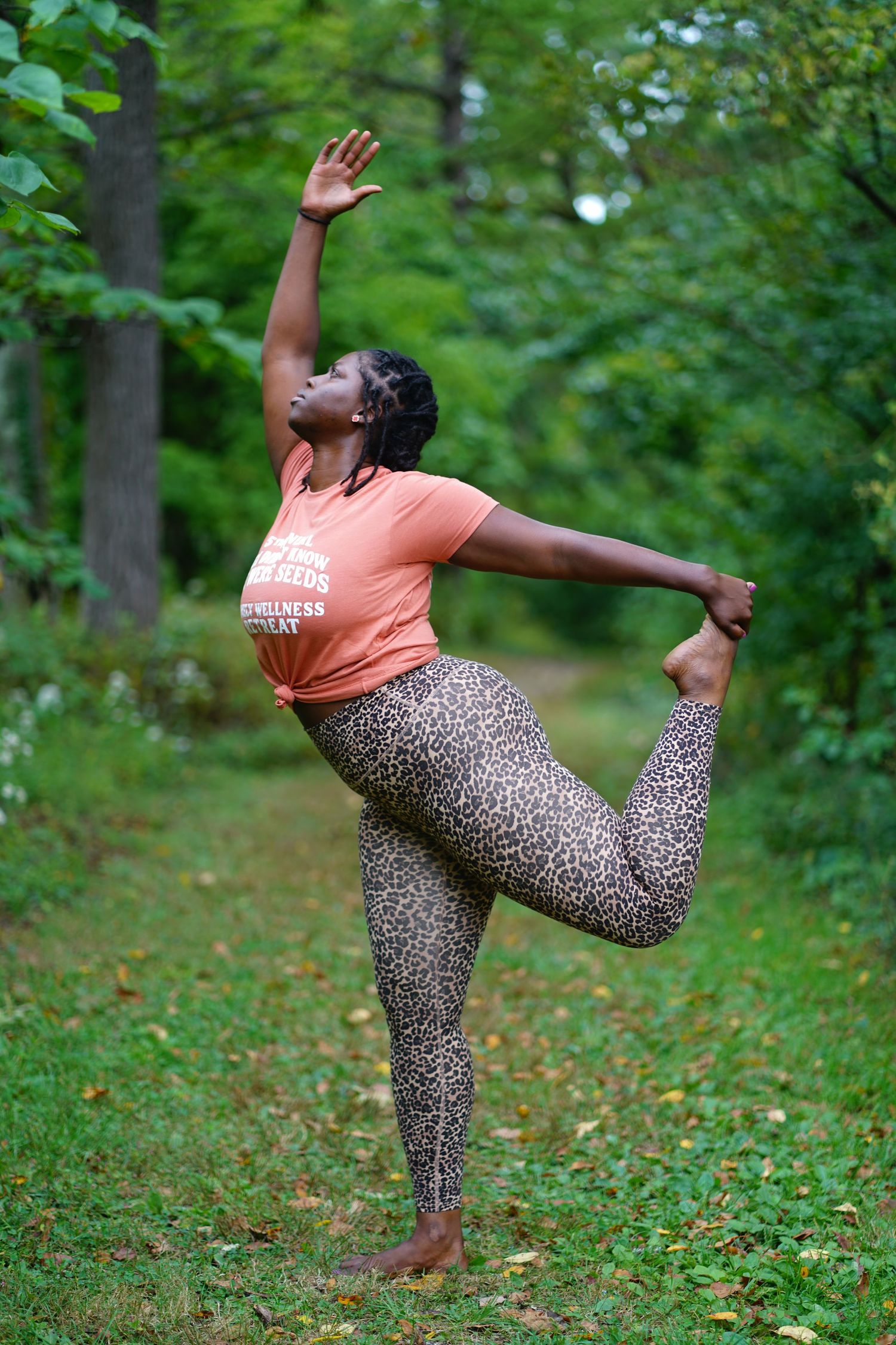 Woman in leopard print leggings and orange top practicing dancer yoga pose outdoors in lush green forest setting.