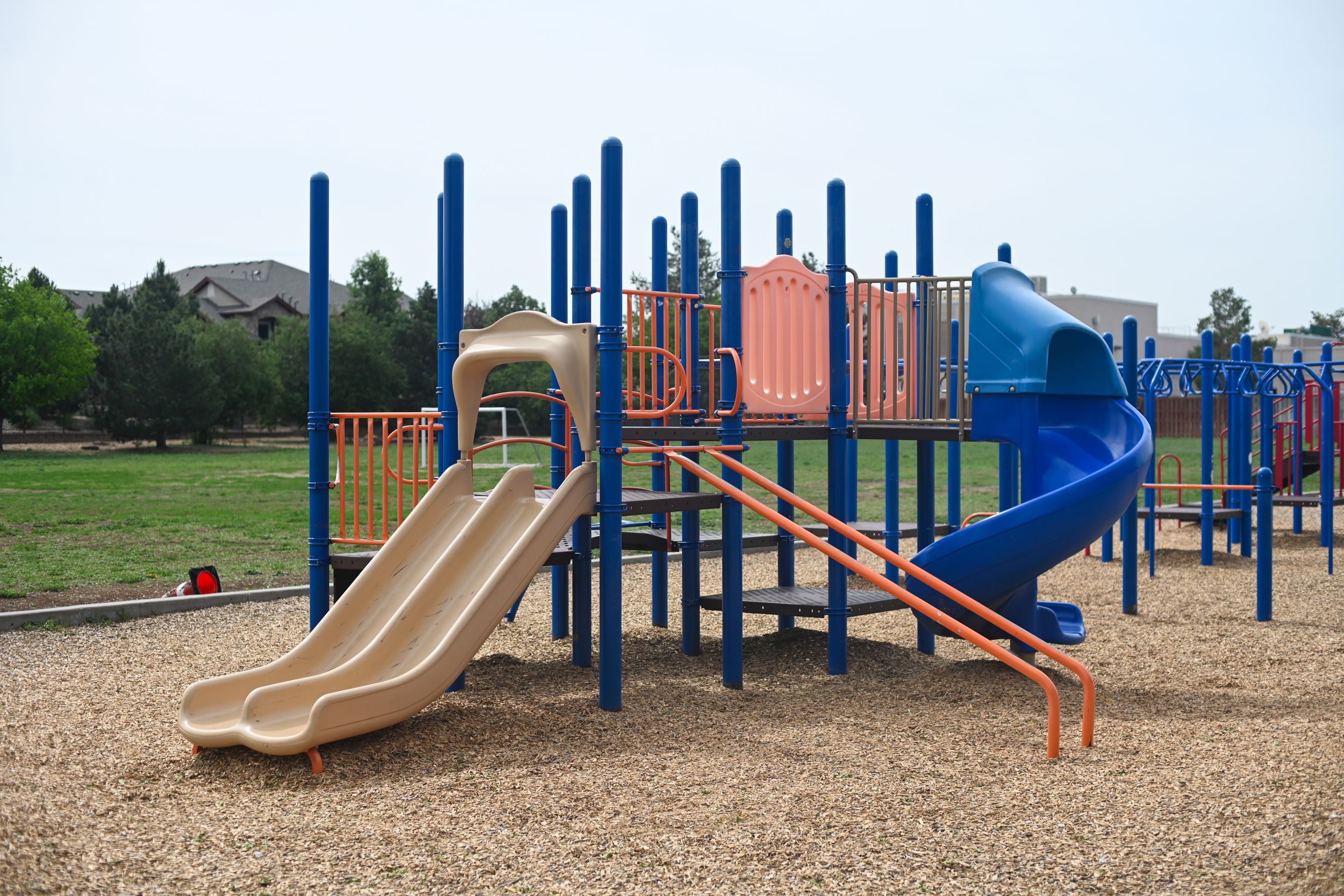 Colorful playground with blue metal frame, dual tan slides, spiral blue slide, and wood chip ground cover in suburban park.