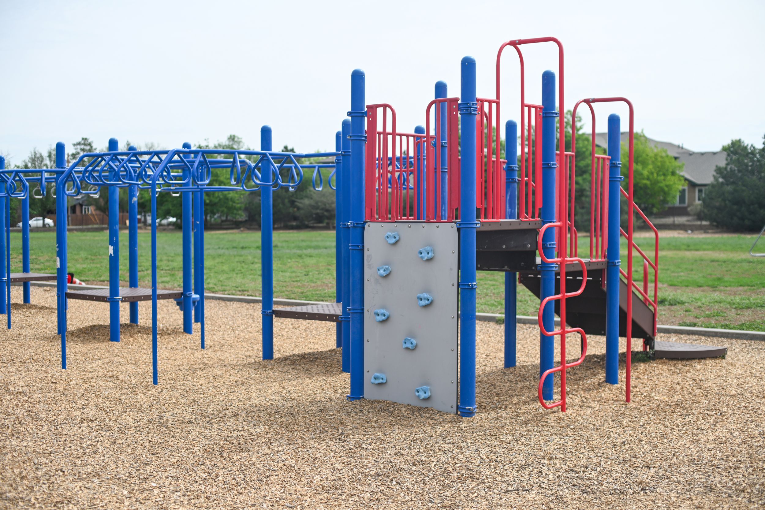 Colorful blue and red playground equipment with climbing structures on wood chip ground in a sunny park.