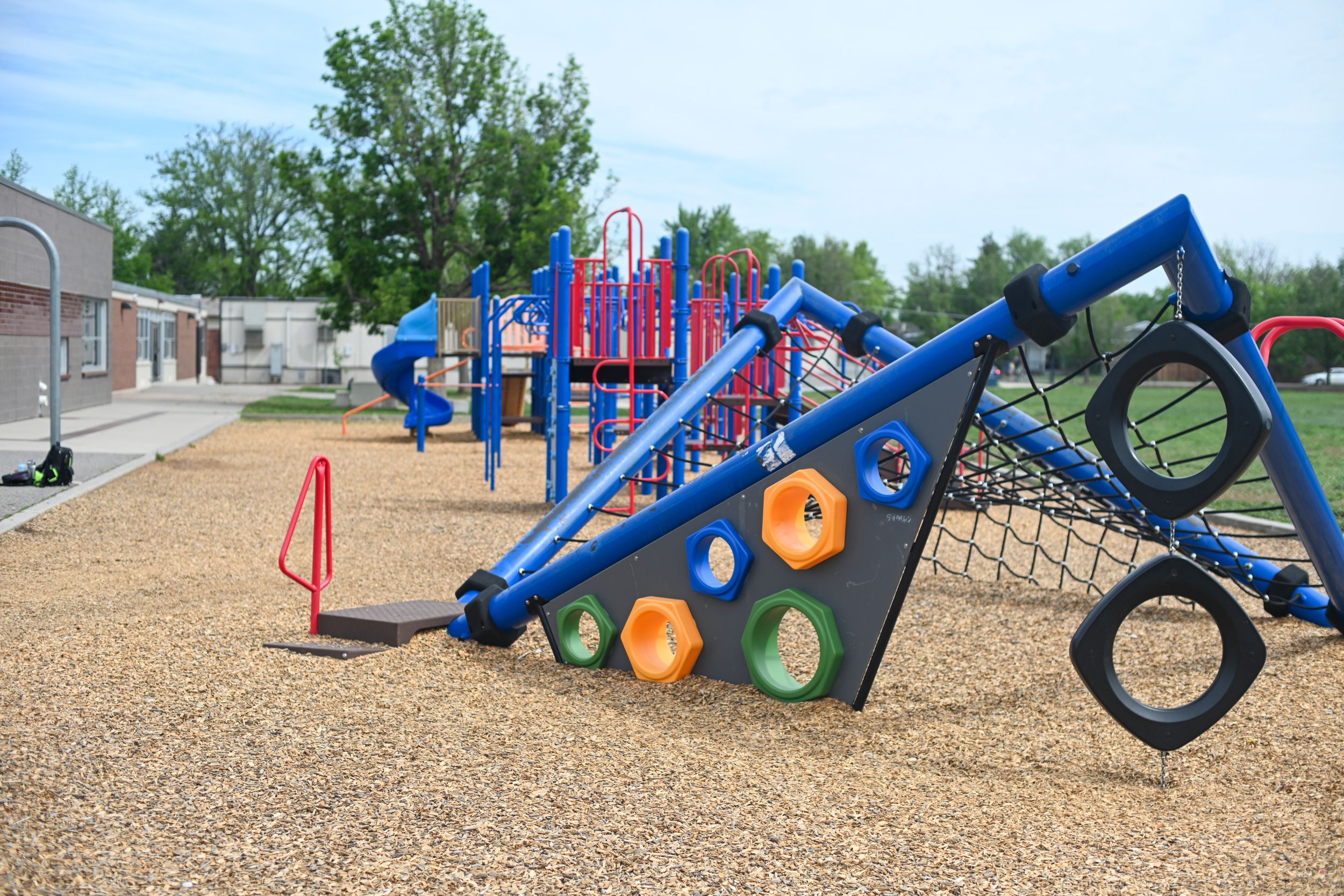 Colorful playground equipment with climbing net, tire swing, and slides on wood chip ground at a school recess area.