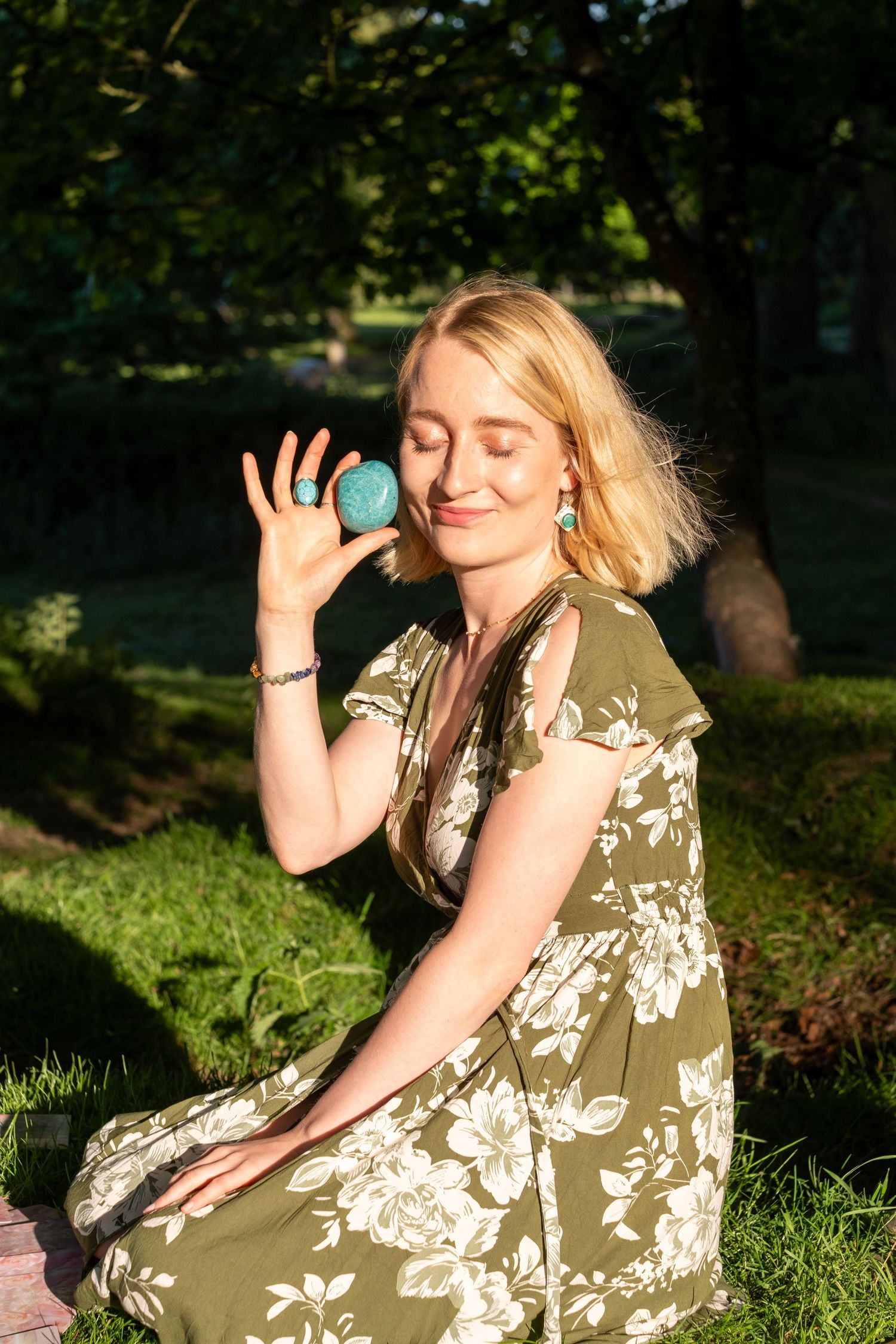 Blonde woman in floral green dress holding turquoise stone with eyes closed in sunny park setting.