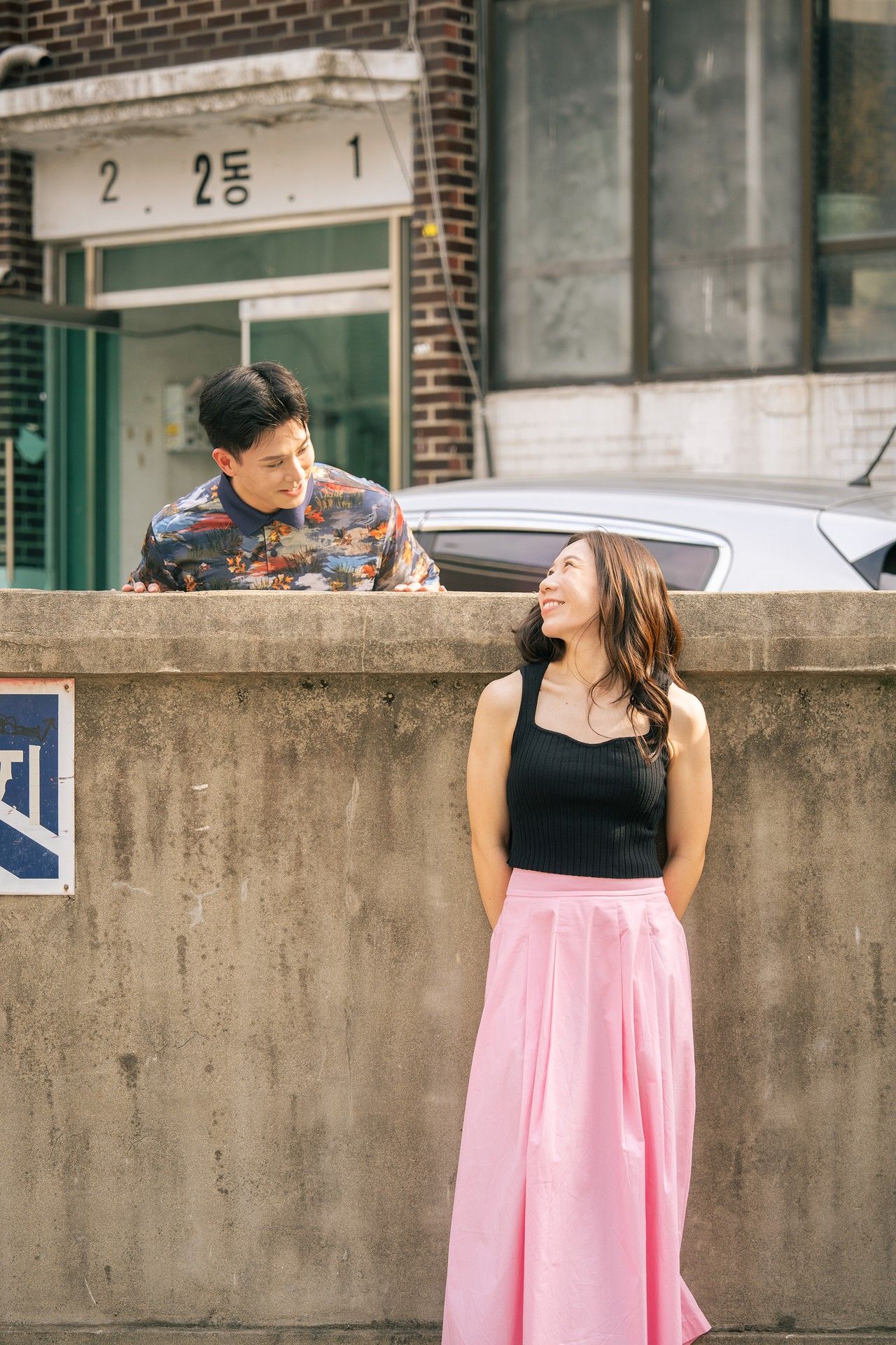 A young couple shares a romantic moment on a Korean street, with the woman in a pink skirt smiling up at the man.