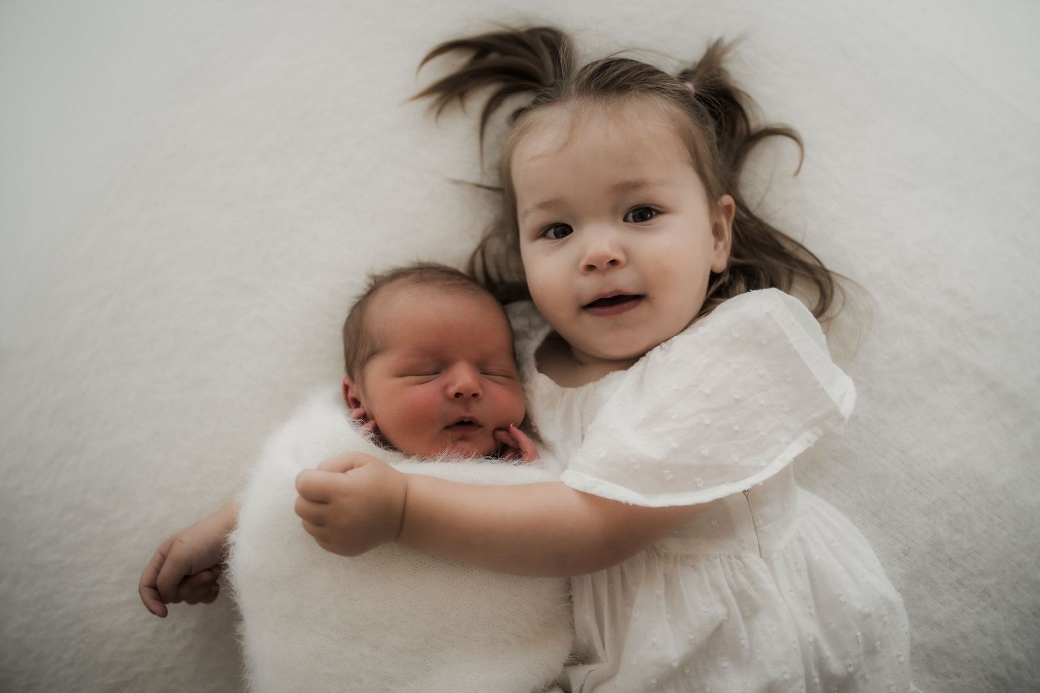 A toddler girl in a white dress lovingly hugs a sleeping newborn baby wrapped in white on a soft blanket in the Buxton, Victoria Studio