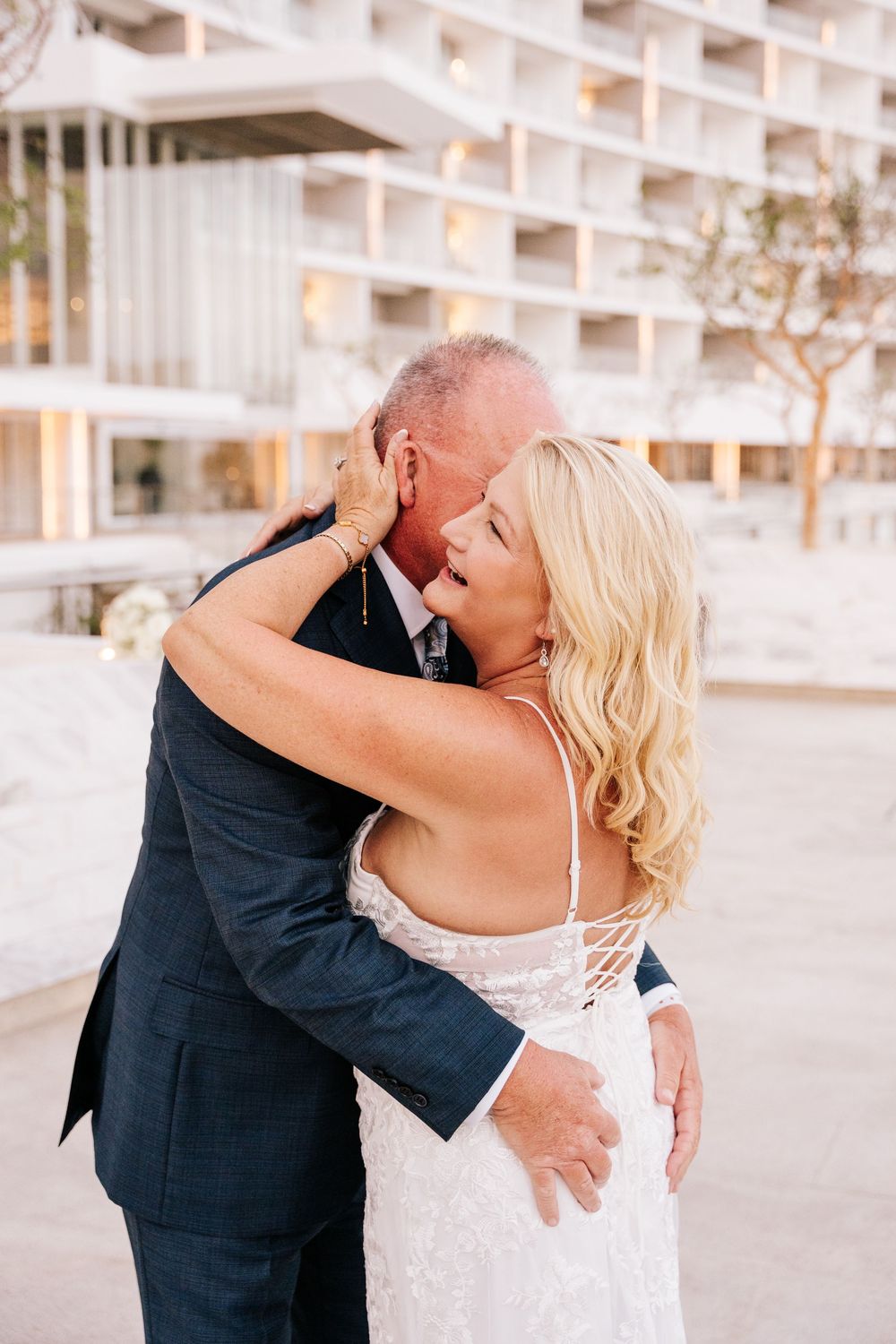 Bride and groom share a romantic embrace outdoors, with the bride in a lace wedding gown and groom in a navy suit.