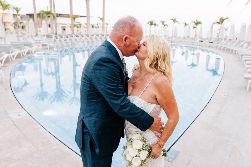 Bride and groom sharing a kiss by a heart-shaped pool, bride holding white bouquet.
