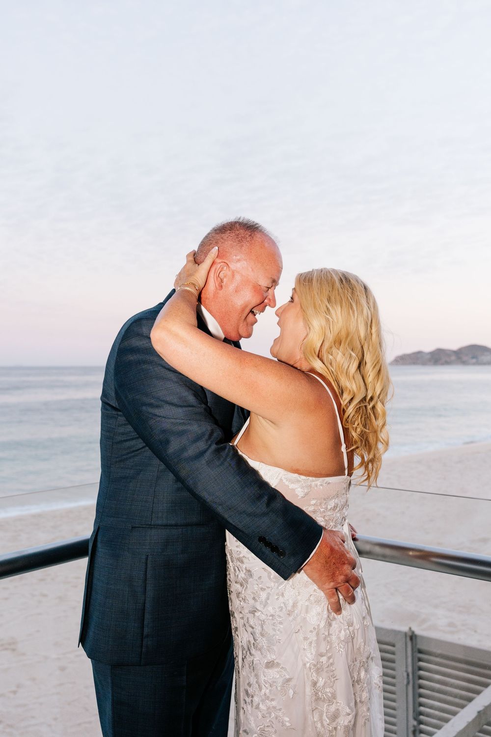 Couple sharing a romantic embrace at sunset on a beach terrace, bride in white lace gown and groom in dark suit.