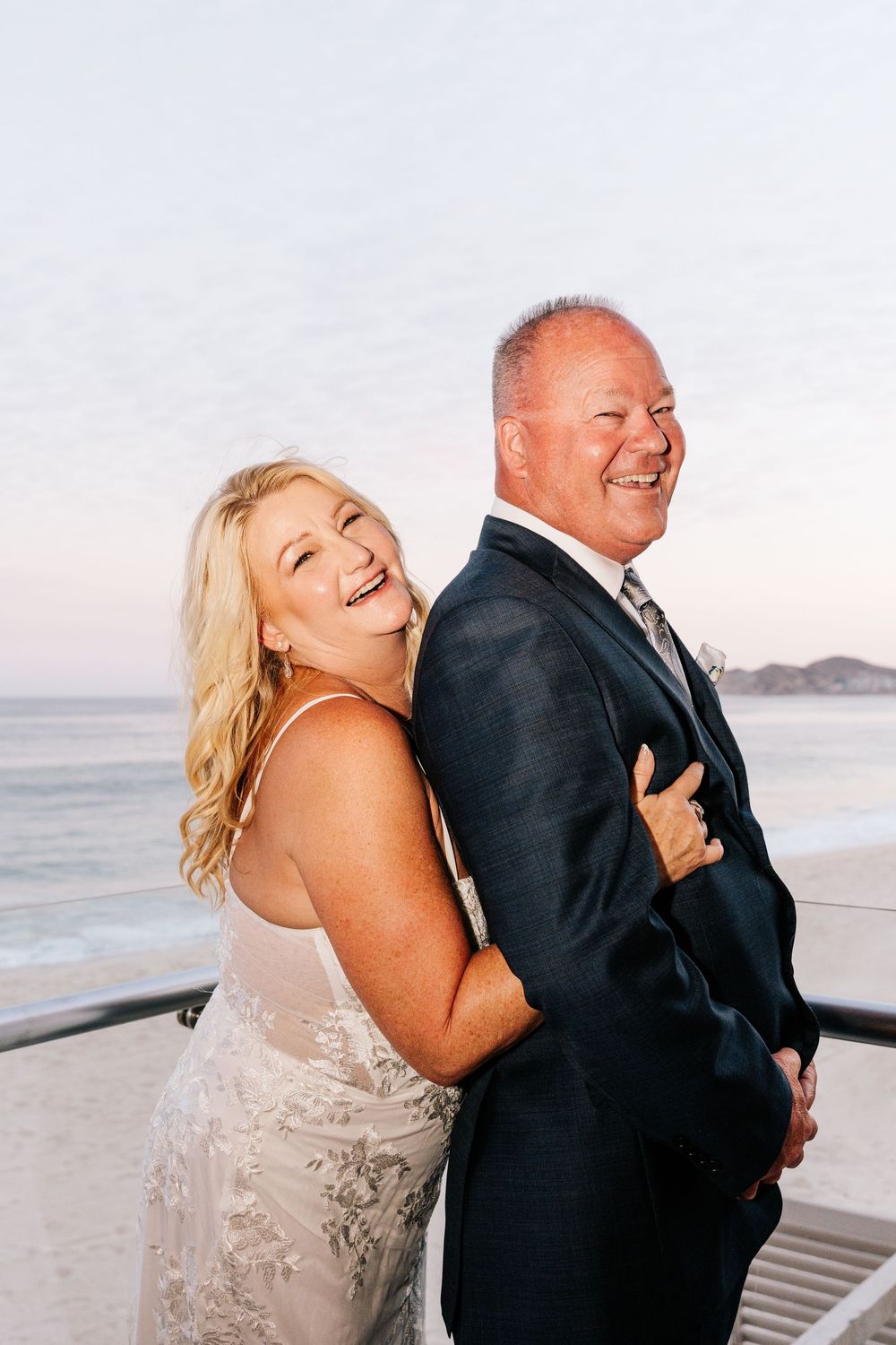 Happy couple in wedding attire laughing together on a beachfront deck at sunset.