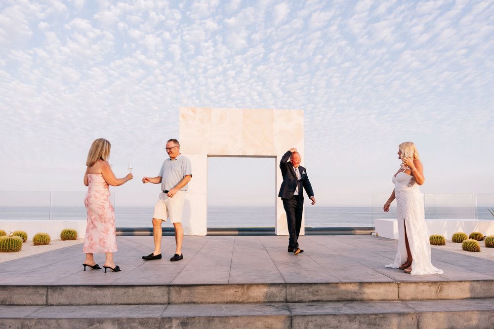 Four people gather at an elegant outdoor terrace with a white arch, ocean view, and dramatic cloudy sky at golden hour.