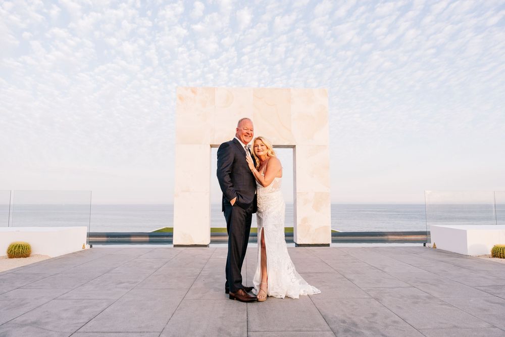 Bride and groom pose under a stone arch at an outdoor oceanview wedding ceremony at sunset.