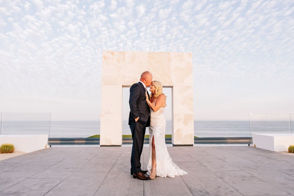 Couple sharing a kiss at outdoor wedding ceremony with ocean backdrop and decorative arch at sunset.