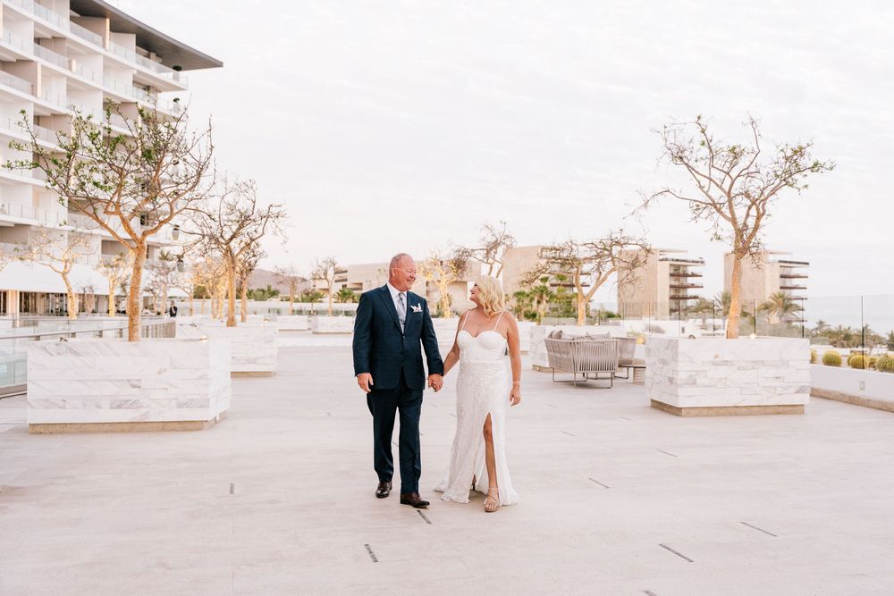 Bride and groom hold hands on a sunlit terrace with modern architecture and bare trees at a destination wedding.