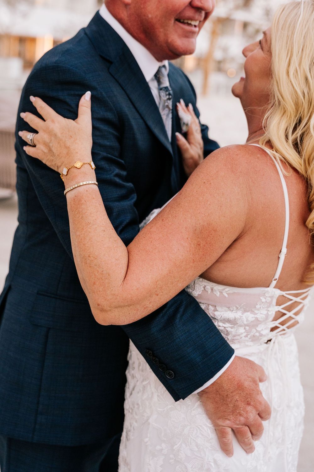 A bride in a lace gown embraces a groom in a navy suit, smiling joyfully during their wedding celebration.