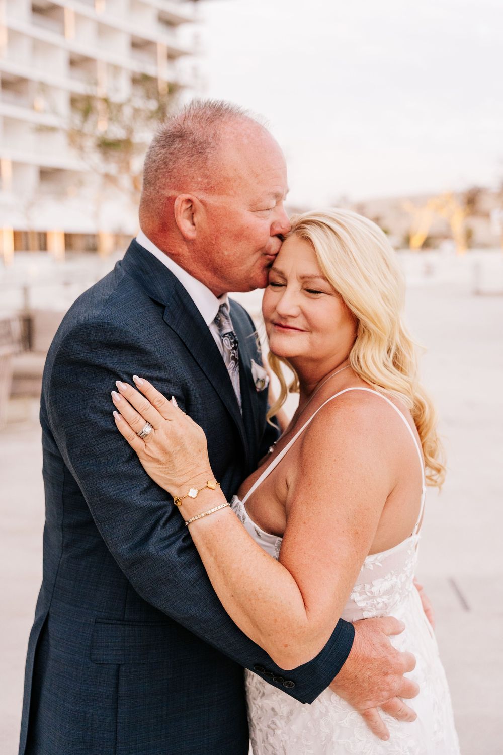 A groom in a navy suit kisses his blonde bride on the cheek as she smiles with eyes closed outdoors.