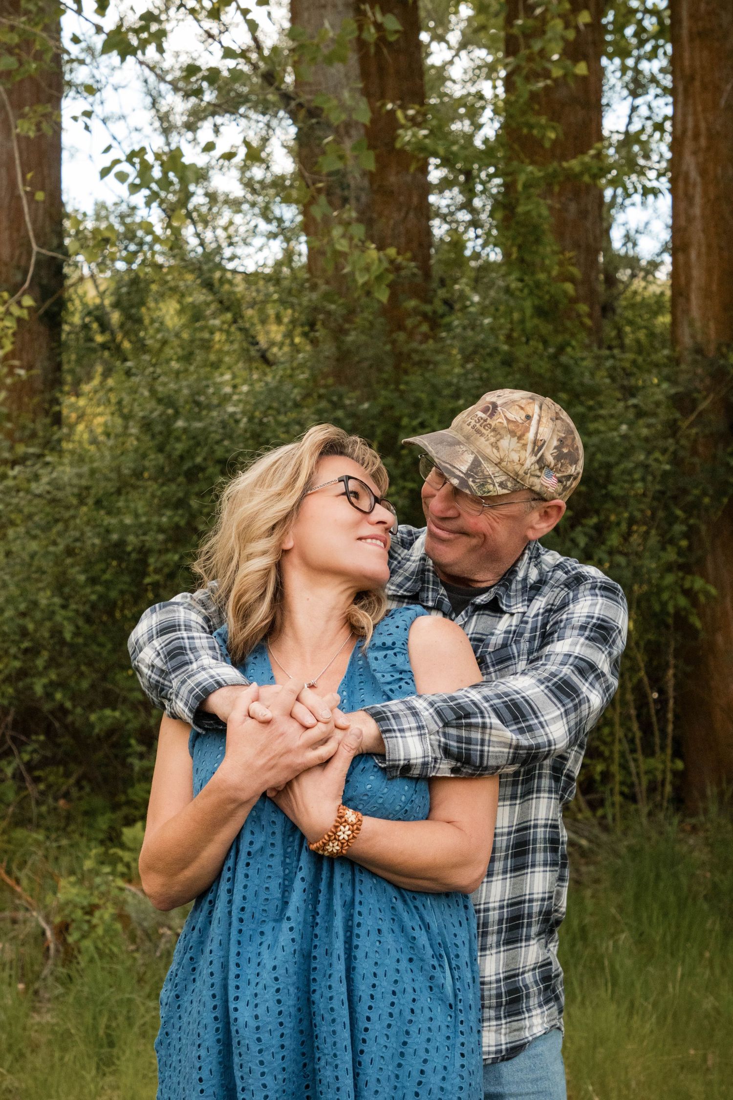 A couple embraces outdoors in a forest, smiling at each other, woman in blue dress, man in plaid shirt and camo cap.