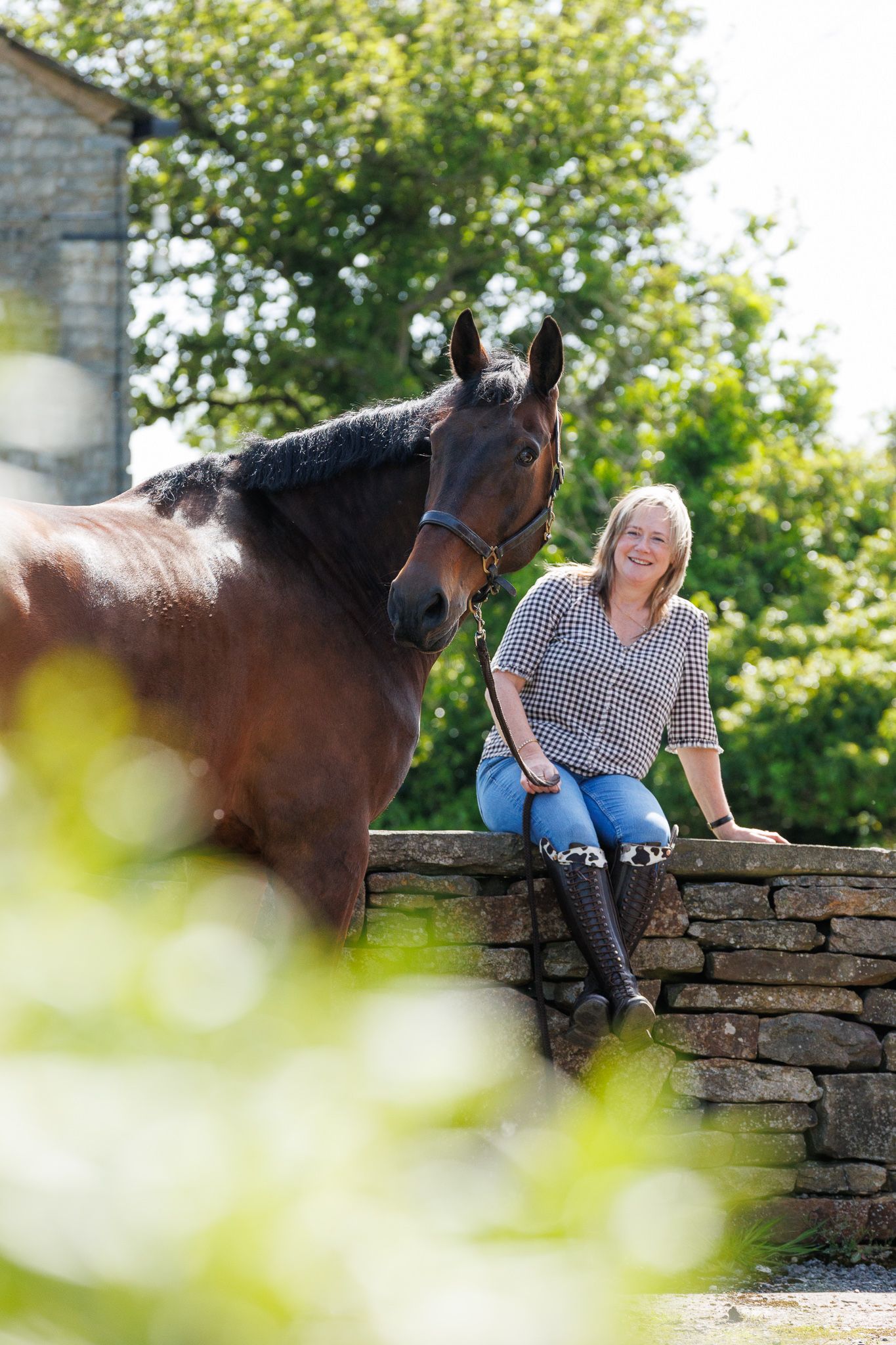 Blonde woman in jeans and boots sits on a stone wall beside a bay horse outdoors.
