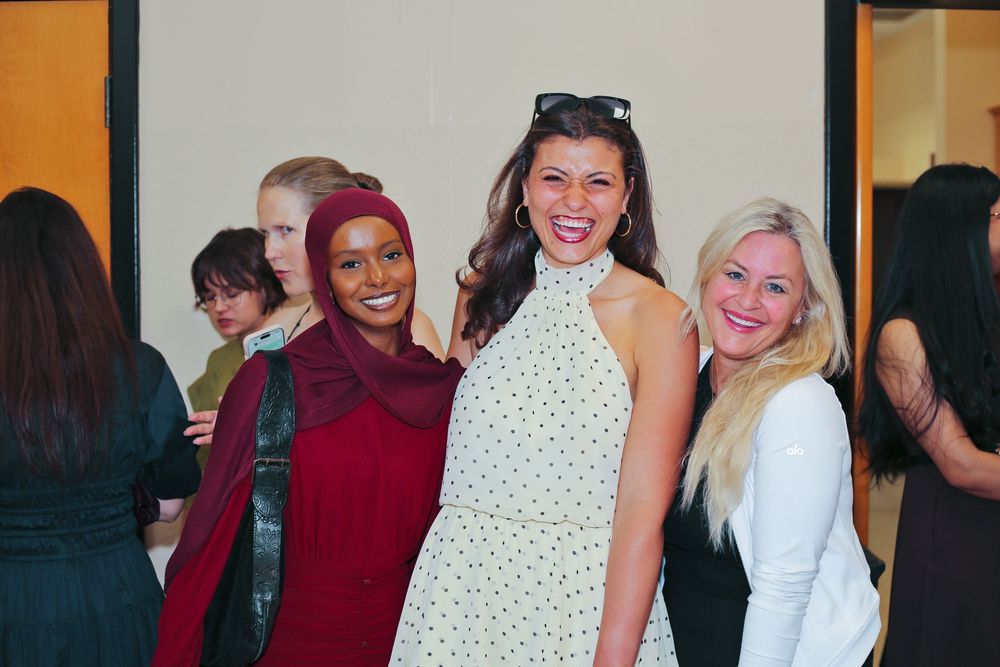 Three smiling women pose together at a social gathering, dressed in elegant attire including red hijab and white dresses.