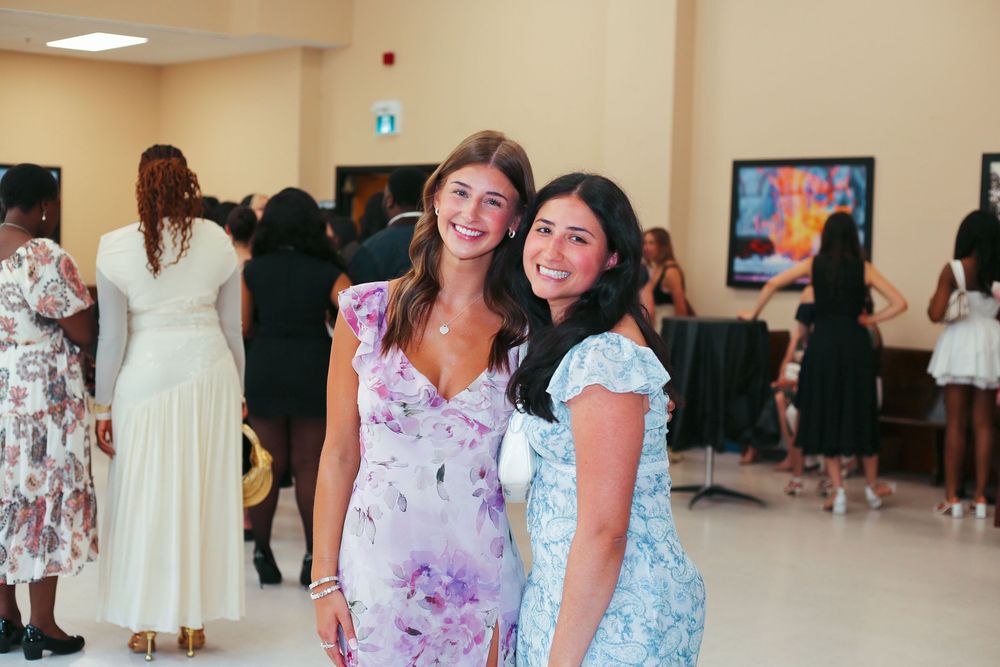 Two smiling women in floral dresses pose together at an elegant indoor event with guests in background.
