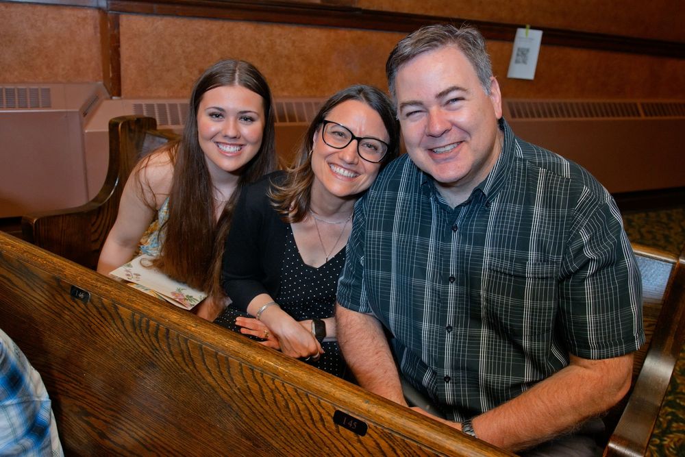 Family of three smiling together in wooden church pews, posing for a warm portrait photo.