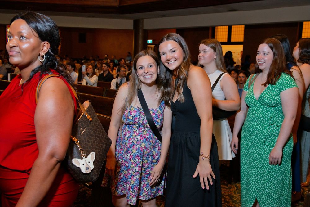 Women smiling and posing together at a formal indoor event in a crowded auditorium.