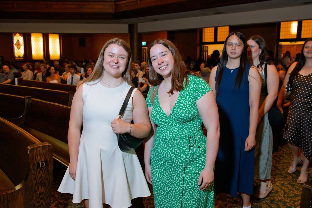 Two young women in dresses smiling together at a formal indoor ceremony or event.