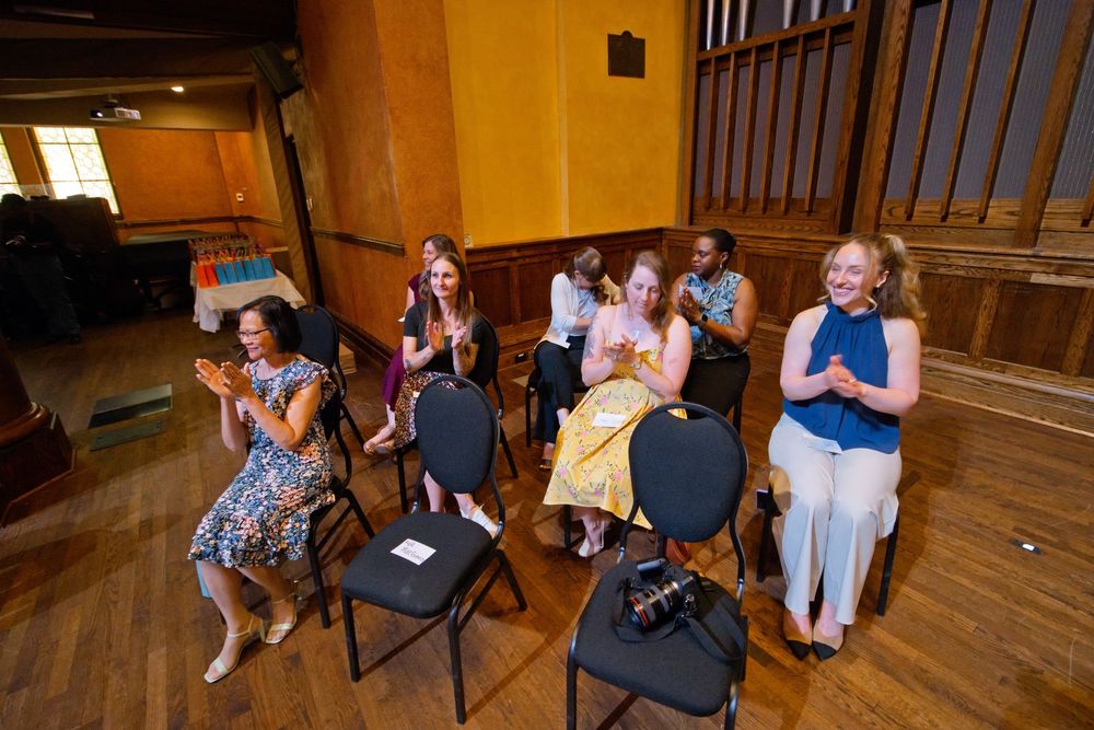 Group of women clapping and smiling while seated on folding chairs in a warmly lit wooden hall.