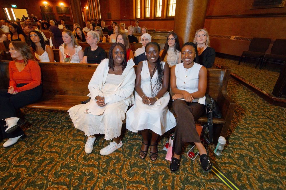 Three women smile while seated in ornate theater pews, with a diverse audience gathered in rows behind them.