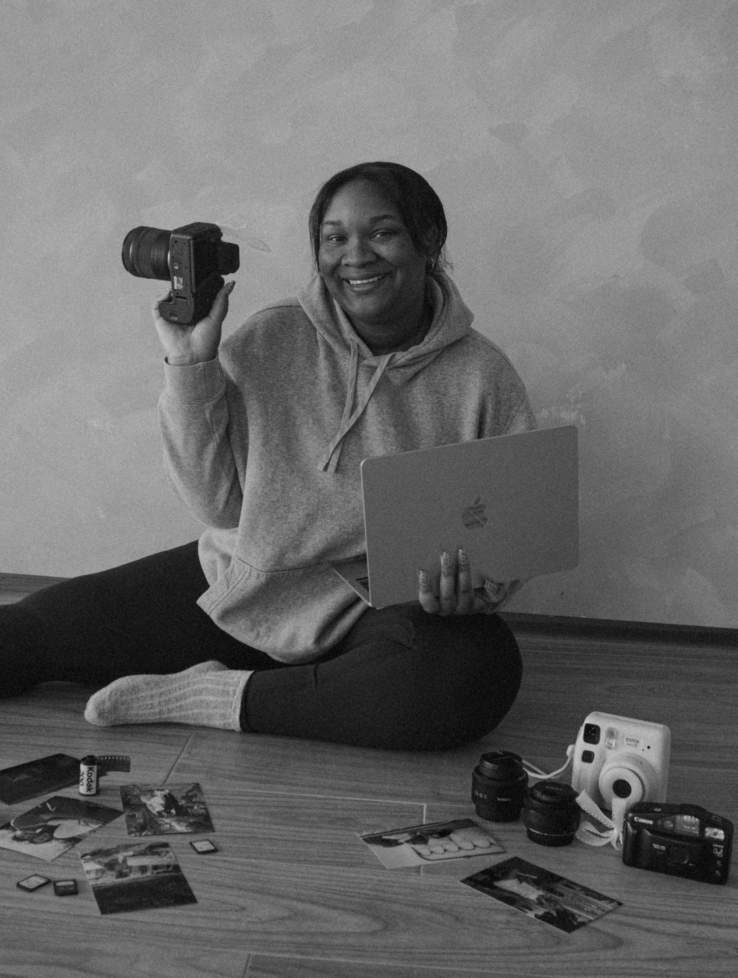 Smiling woman sitting on floor holding DSLR camera and Apple laptop surrounded by photos and cameras in black and white photo.