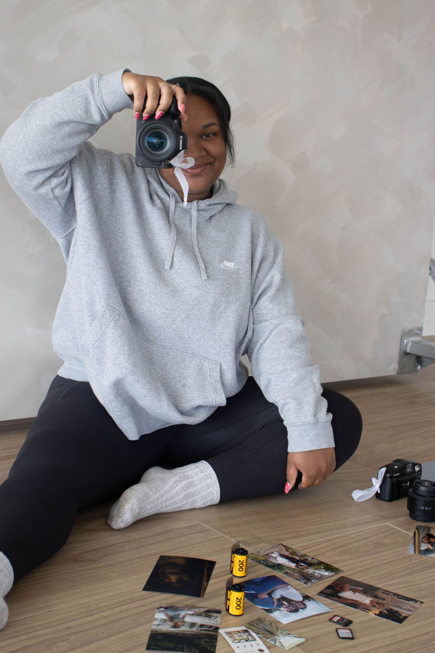 Woman in gray hoodie sits on floor, holding DSLR camera up to take photo, surrounded by printed photos and film rolls.