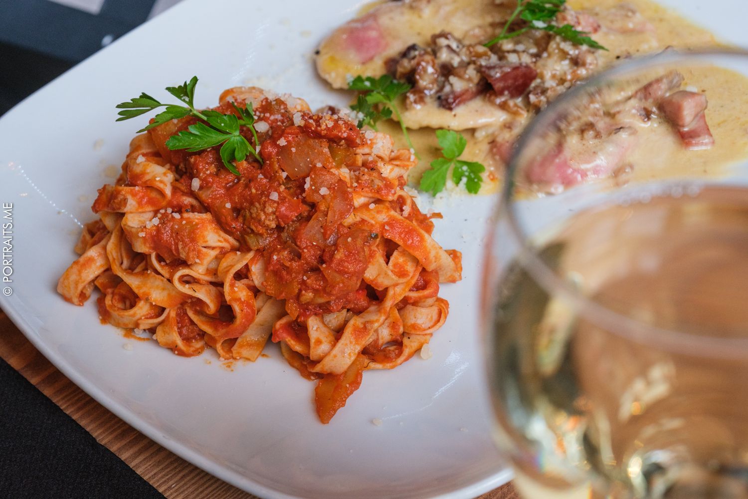 A white plate with fettuccine bolognese and a creamy pasta dish, garnished with parsley, beside a glass of white wine.