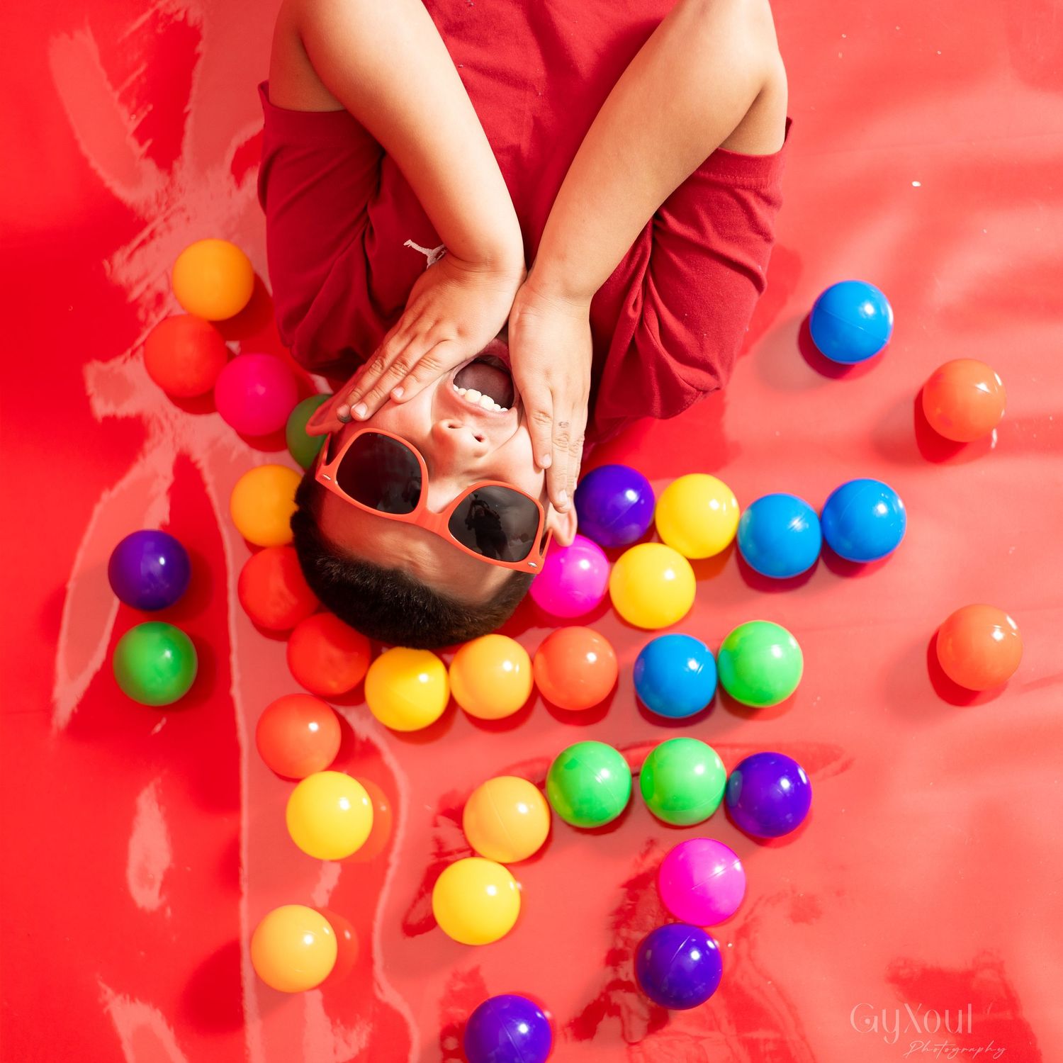Child in red outfit lying on red surface surrounded by colorful plastic balls, wearing red sunglasses, top-down view.