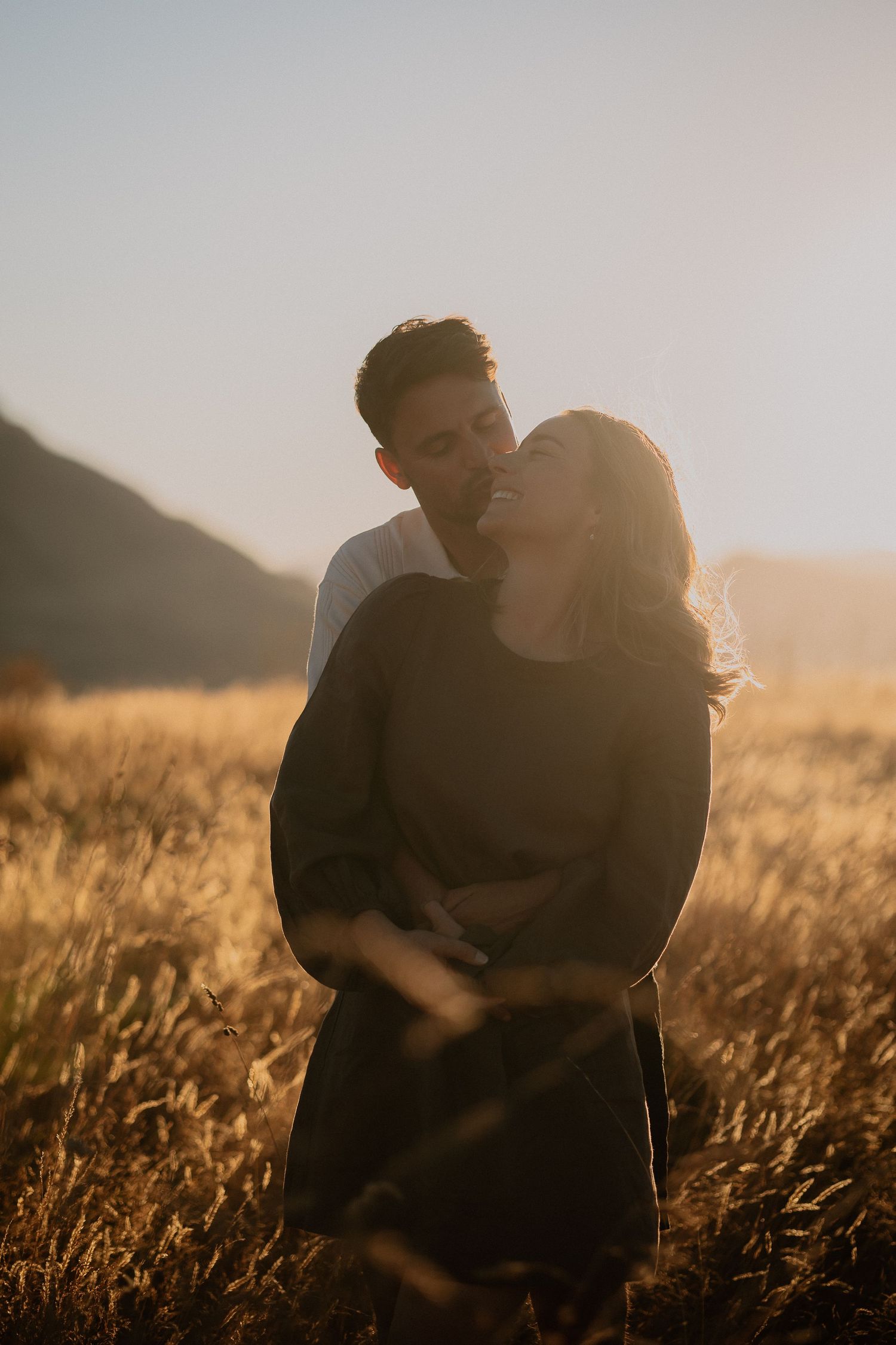 Couple embracing in golden sunlit field with tall grass in foreground during sunset.