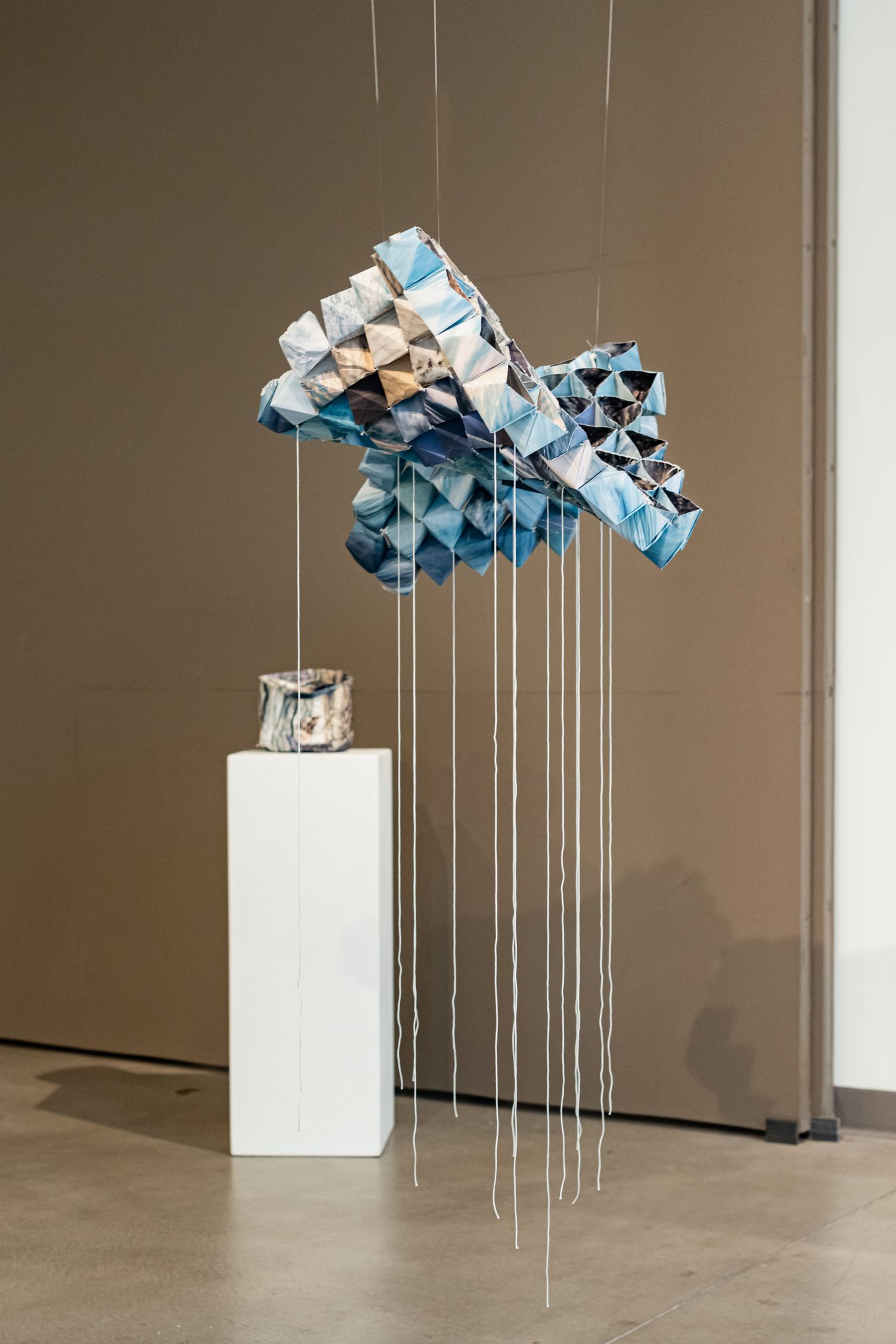 A geometric origami sculpture in blue tones suspended by strings above a white pedestal in a gallery.