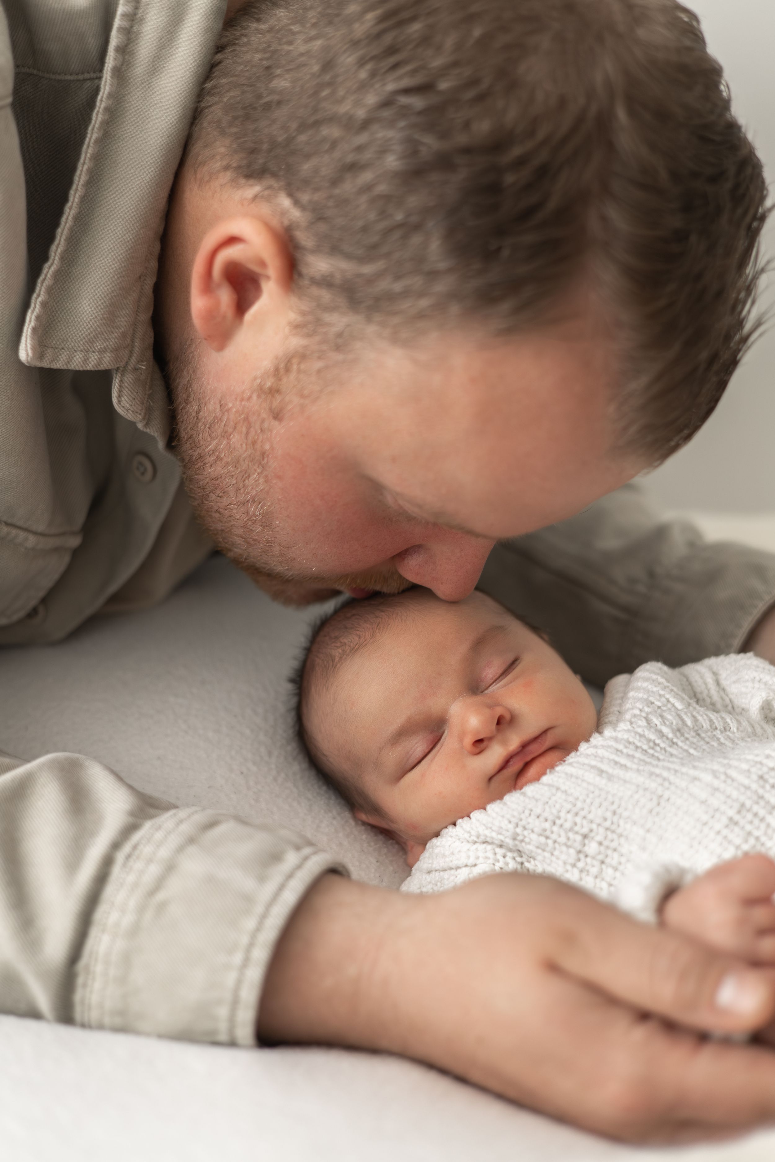 A father gently kisses his sleeping newborn baby wrapped in white, sharing a tender moment. during natural Newborn photoshoot Nottingham