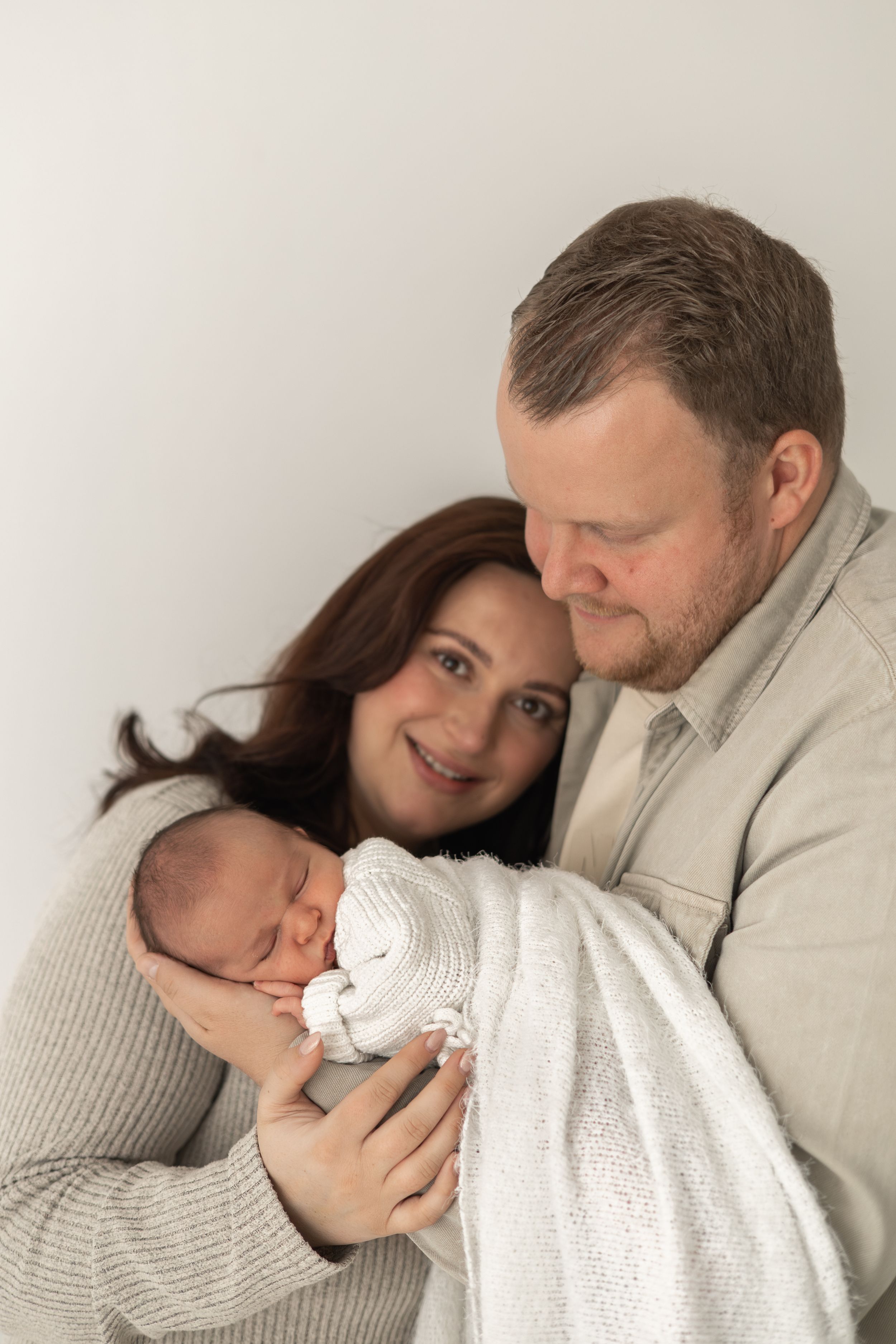 A mum and dad smiles lovingly at their sleeping newborn baby wrapped in a white blanket during natural Newborn photoshoot Nottingham