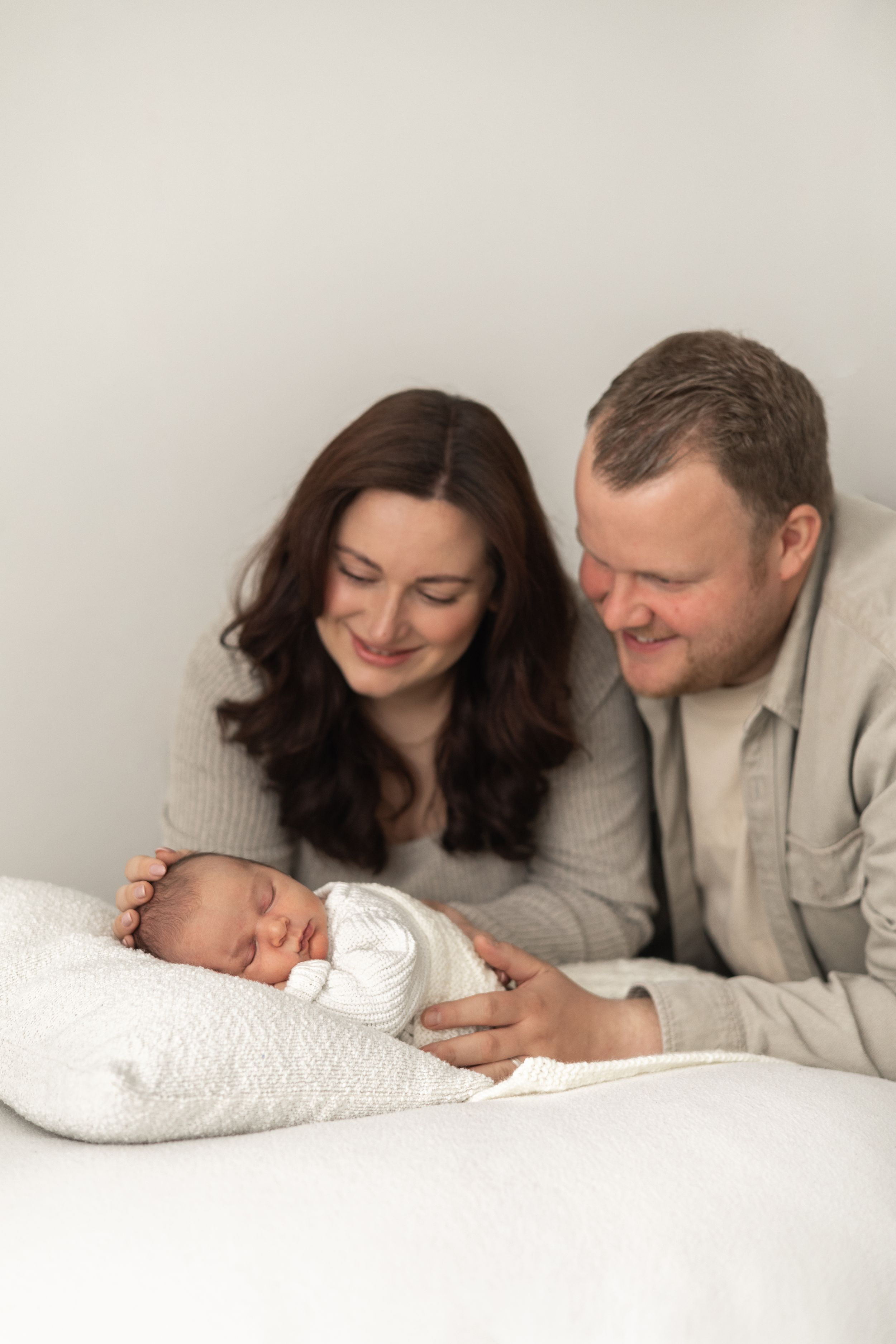 A mum and dad gazes lovingly at their sleeping newborn baby wrapped in white during natural Newborn photoshoot Nottingham