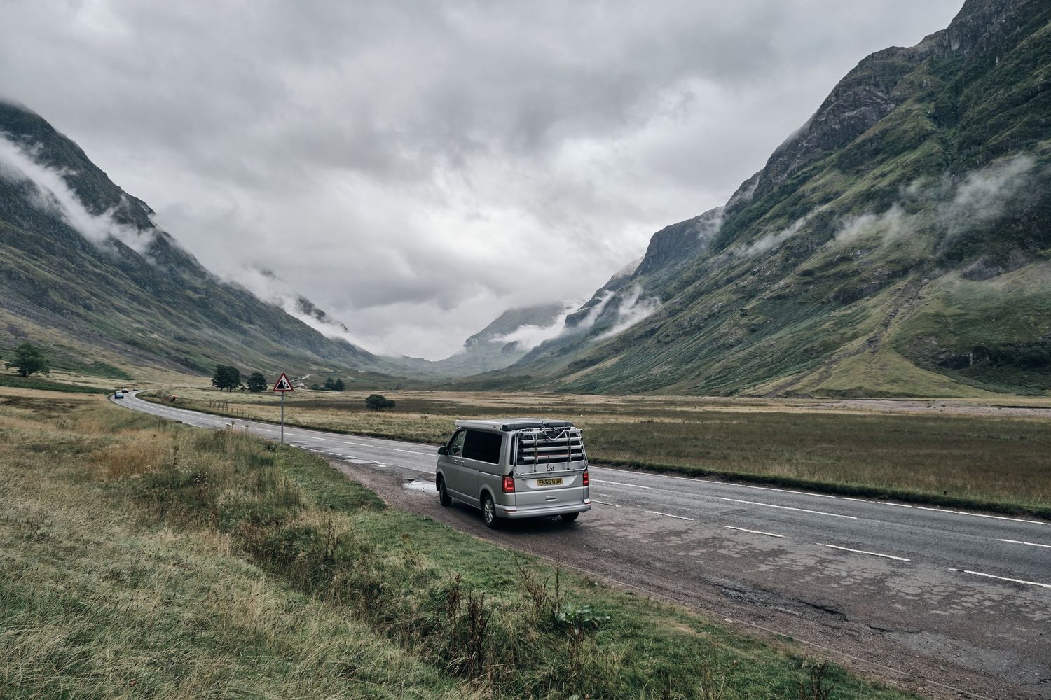 A silver van drives through a dramatic Scottish Highland valley with misty mountains and overcast skies.