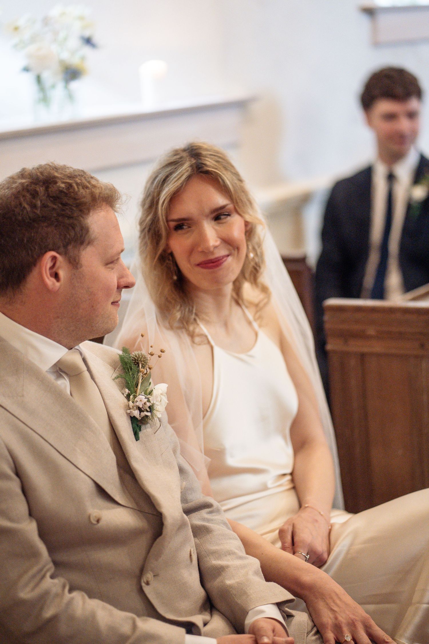 Bride in white dress and veil smiles at groom in tan suit during wedding ceremony in a church.