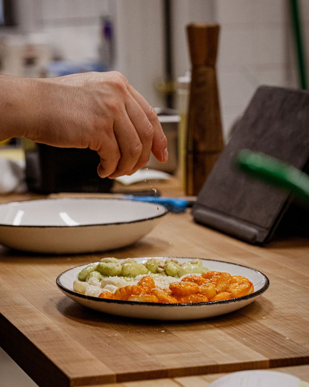 Chef sprinkling seasoning over a tricolor gnocchi dish on a wooden kitchen counter.