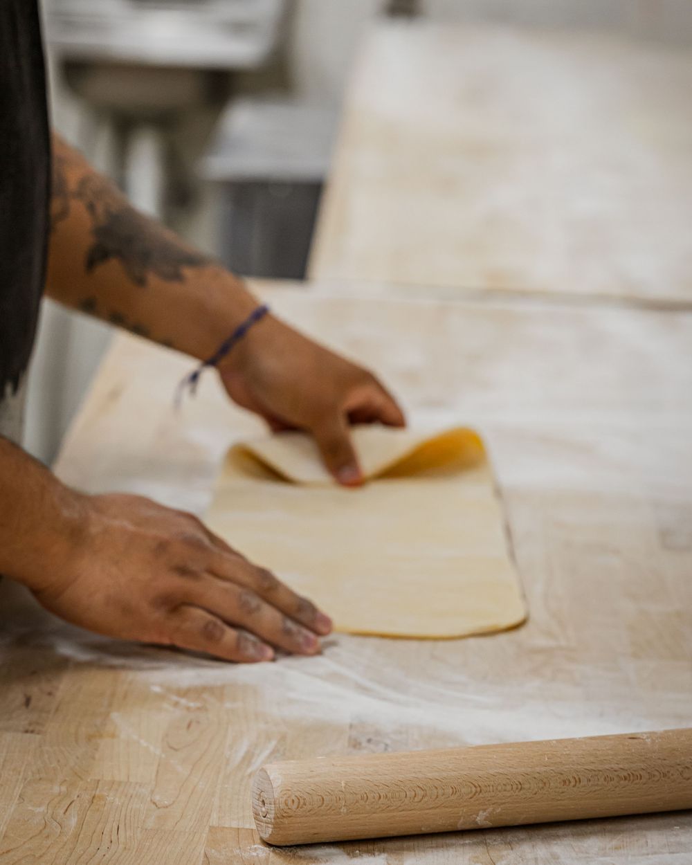 Tattooed hands rolling fresh pasta dough on a flour-dusted wooden surface with a rolling pin nearby.