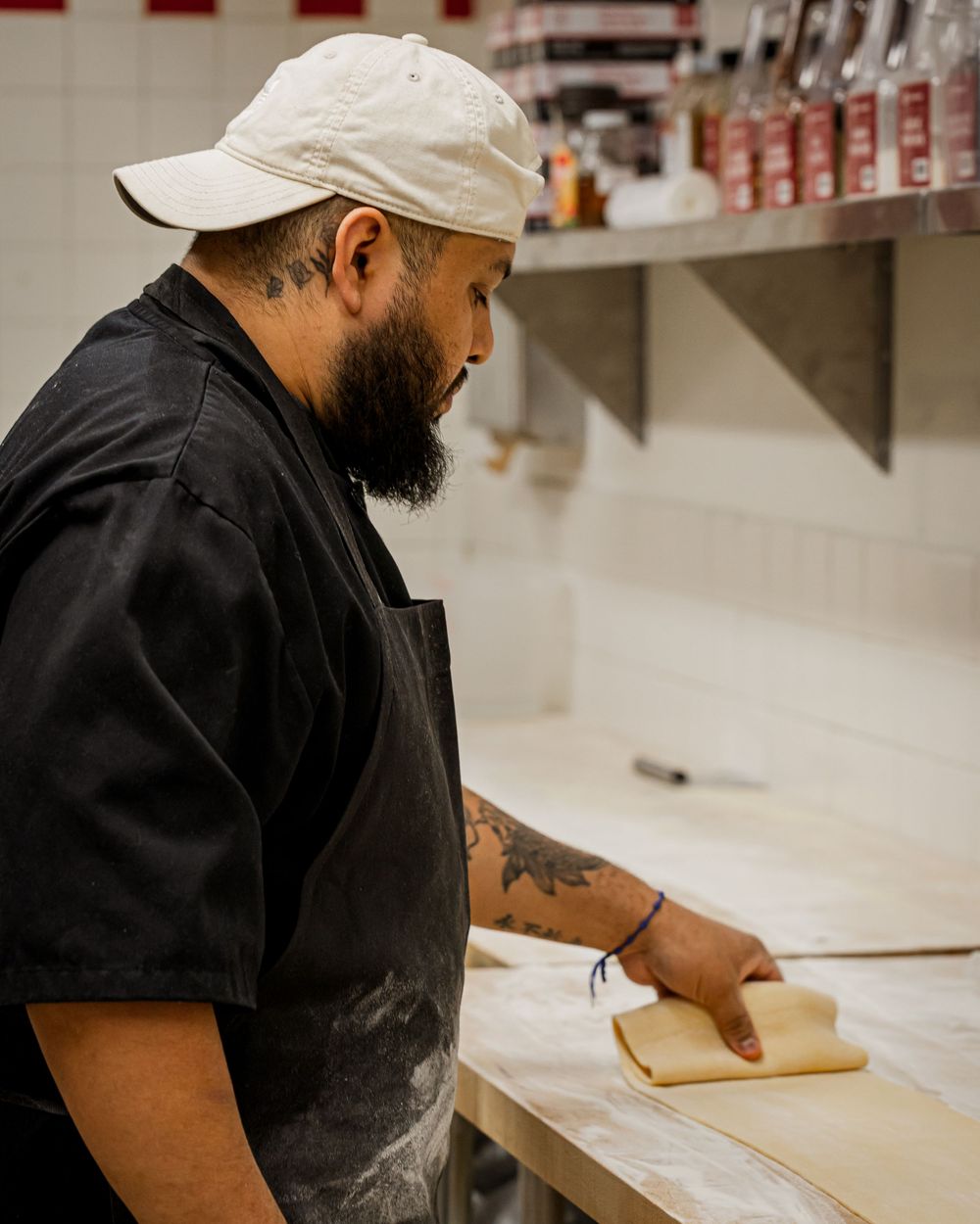 Bearded chef in black uniform and white cap working with fresh pasta dough on a floured wooden surface.