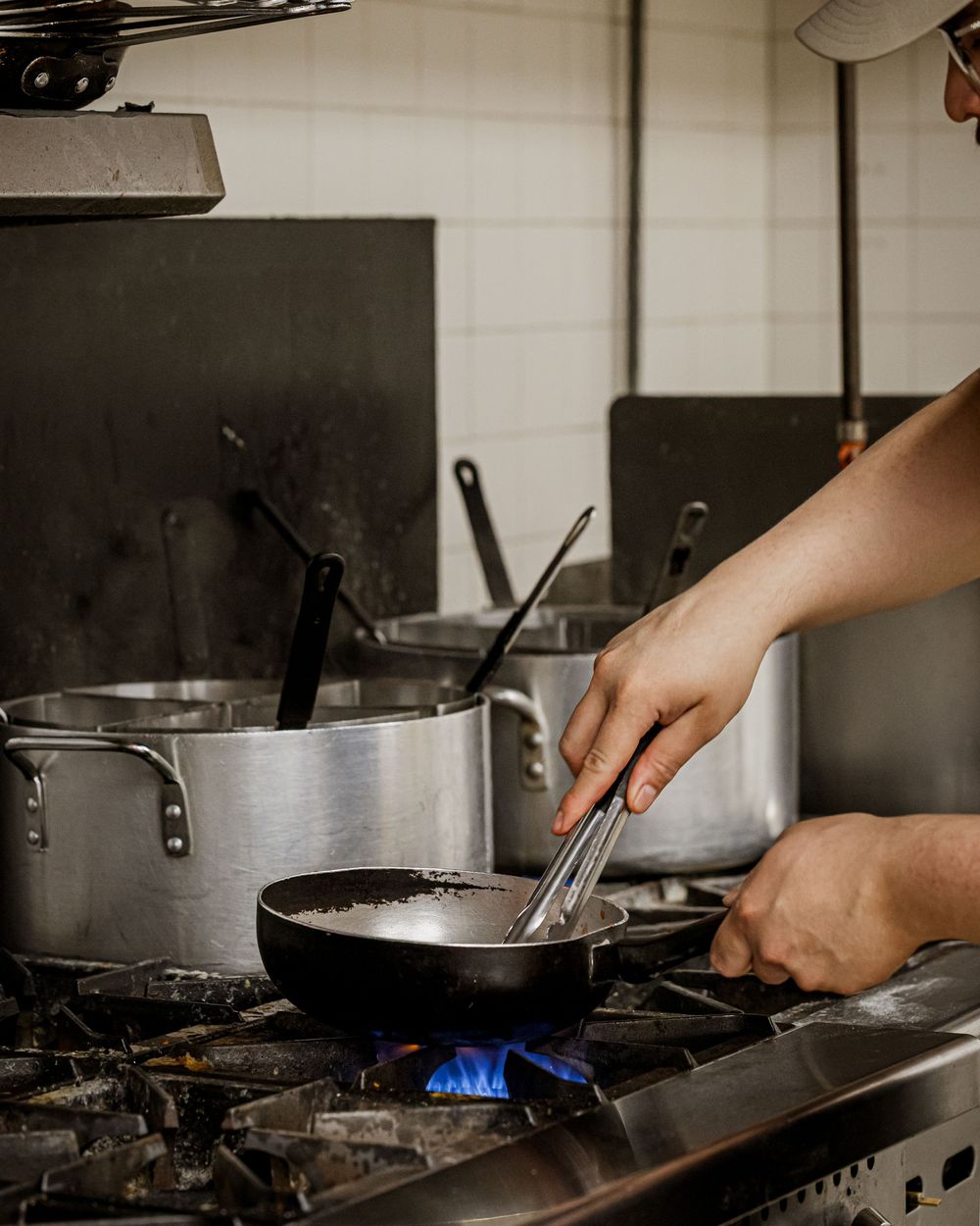 Chef cooking with tongs in a professional kitchen over a gas flame with stainless steel pots.