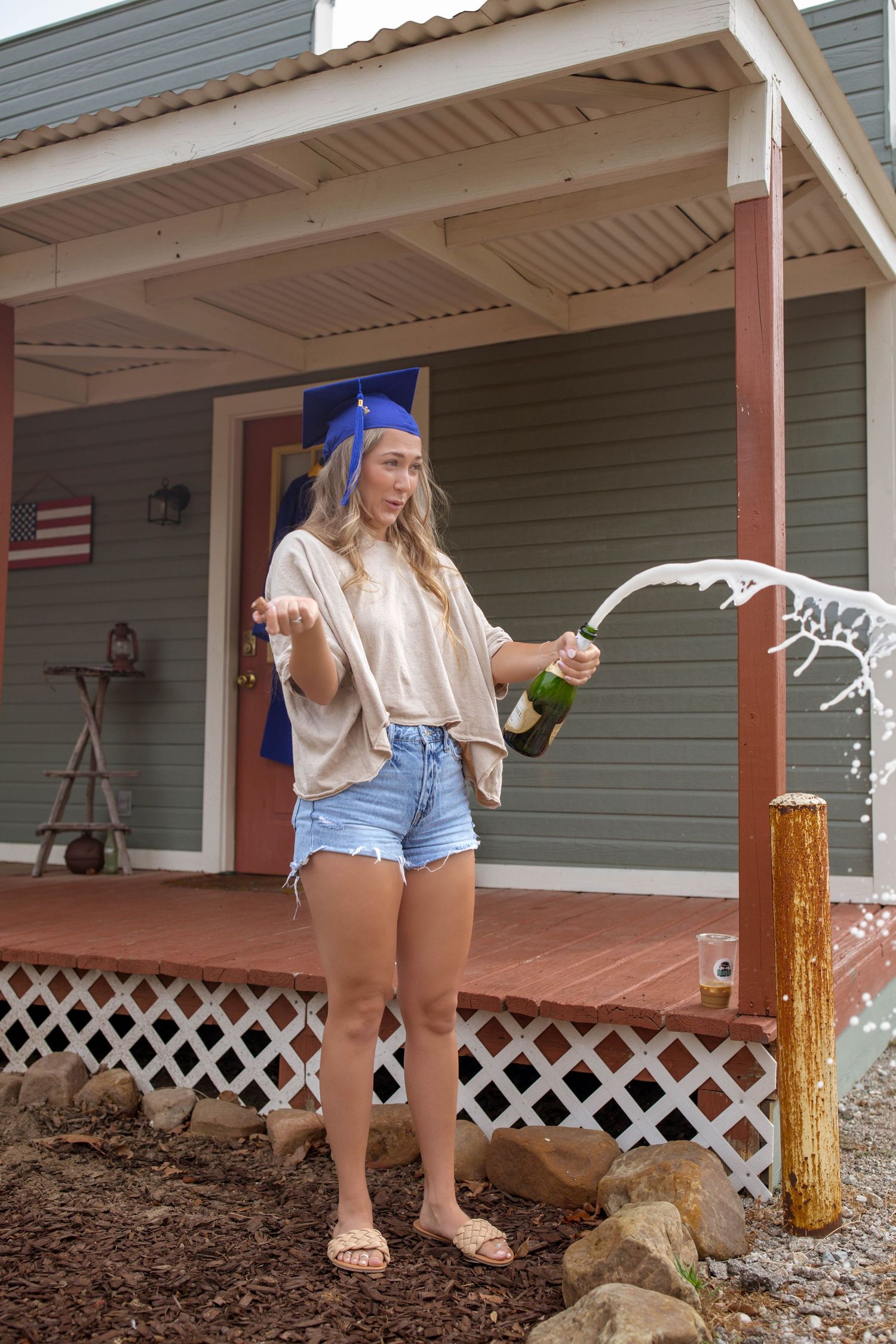Female graduate in blue cap celebrating graduation by popping a champagne bottle outdoors.